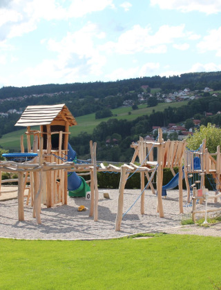 Parque infantil de madera en Camping Lac des Brenets, parque de vacaciones en Neuchâtel, Suiza, con colinas al fondo.