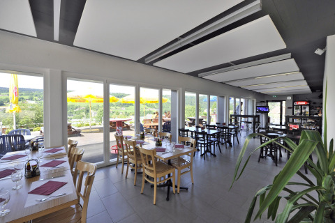 Indoor restaurant with floor-to-ceiling windows, terrace and scenic hill views at Camping Lac des Brenets.