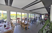 Indoor restaurant with floor-to-ceiling windows, terrace and scenic hill views at Camping Lac des Brenets.