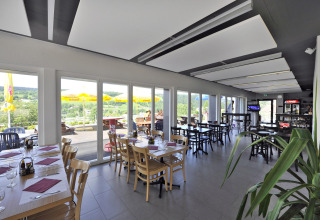Indoor restaurant with floor-to-ceiling windows, terrace and scenic hill views at Camping Lac des Brenets.