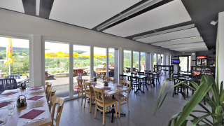 Restaurante interior con ventanas panorámicas, terraza y vistas a colinas en Camping Lac des Brenets.
