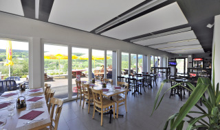 Restaurante interior con ventanas panorámicas, terraza y vistas a colinas en Camping Lac des Brenets.