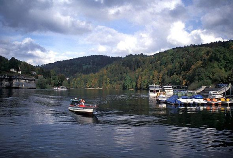 View of the lake at Camping Lac des Brenets in Neuchâtel, Switzerland, with boats, docks, and forested hills.