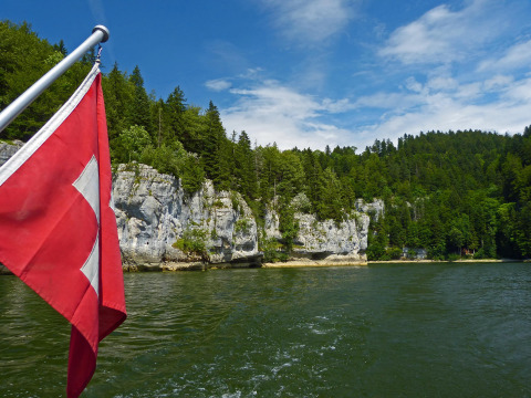 View of Lake at Camping Lac des Brenets in Neuchâtel, Switzerland, with Swiss flag and forested limestone cliffs.