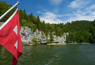 View of Lake at Camping Lac des Brenets in Neuchâtel, Switzerland, with Swiss flag and forested limestone cliffs.
