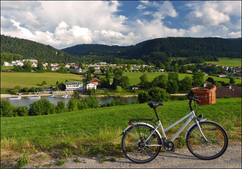 Vista su villaggio, lago, barche e colline vicino a Camping Lac des Brenets, Neuchâtel, Svizzera, bici in primo piano.