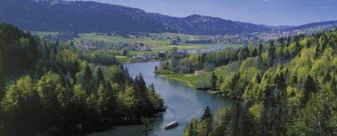 View of a lake surrounded by forest and mountains at Camping Lac des Brenets holiday park in Neuchâtel, Switzerland.