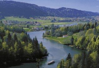 View of a lake surrounded by forest and mountains at Camping Lac des Brenets holiday park in Neuchâtel, Switzerland.