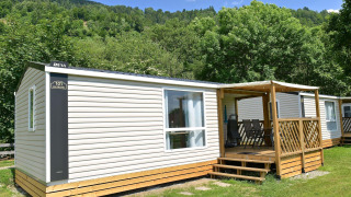 Mobilhome med terrasse i grønne omgivelser, lyst panel og udendørs siddeområde under blå himmel.