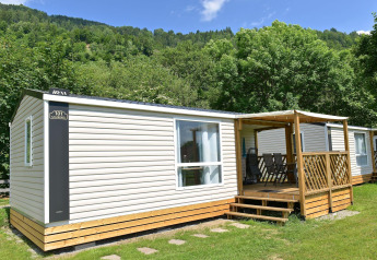 Mobilhome med terrasse i grønne omgivelser, lyst panel og udendørs siddeområde under blå himmel.