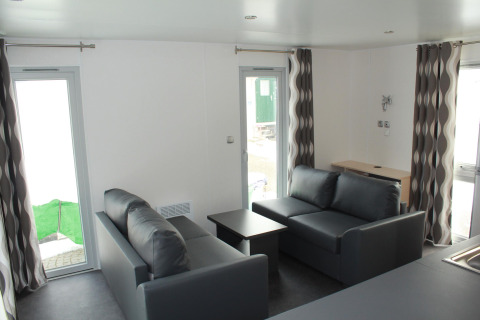 Living area in Mobile home O'Hara at Camping Lac des Brenets, Switzerland, featuring grey sofas and bright windows.
