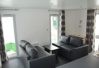 Sala de estar en la casa móvil O'Hara en Camping Lac des Brenets, Suiza, con sofás grises y ventanales amplios.