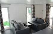 Living area in Mobile home O'Hara at Camping Lac des Brenets, Switzerland, featuring grey sofas and bright windows.
