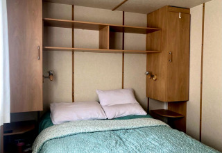 Schlafzimmer im Lavandou Mobilheim auf Camping Lac des Brenets, Schweiz, mit Bett und Regalen.