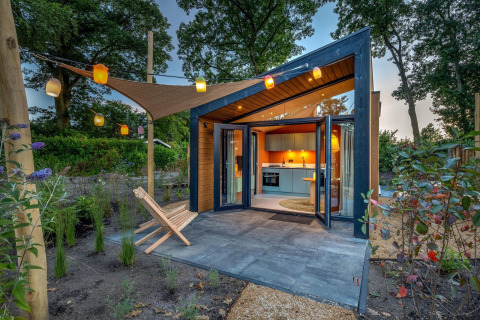 Tiny Wellness Suite, eigentijds tiny house met open glazen deuren, tuin, stoelen en lichte lantaarns bij avond.