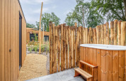 Außenbereich mit Holzbadezuber, rustikalem Zaun und modernen Tiny Houses im Holiday Park De IJsvogel, Niederlande.