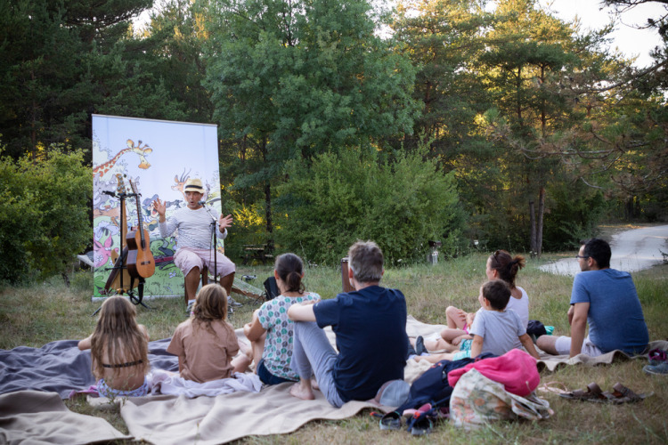 Families and children sit on blankets outdoors listening to a storyteller at Village Huttopia Lagoa de Óbidos.