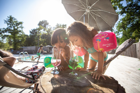 Zwei Kinder mit Schwimmflügeln trinken bunte Getränke am Pool im Village Huttopia Lagoa de Óbidos, Portugal.