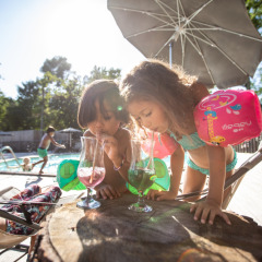 Dos niños con flotadores disfrutan de bebidas coloridas junto a la piscina en Village Huttopia Lagoa de Óbidos, Portugal.