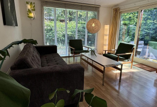 Woonkamer in Retro Lodge bij Bungalowpark Het Verscholen Dorp met grote ramen, retro meubels en groen uitzicht.