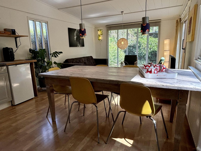 Woonruimte met houten tafel, gele stoelen en grote ramen in Retro Lodge, Bungalowpark Het Verscholen Dorp, Nederland.