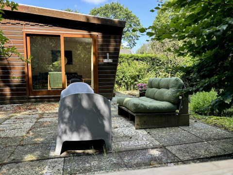 Buitenzitje aan Retro Lodge met groene tuinzetel, stoel en zicht op houten chalet en groene tuin.