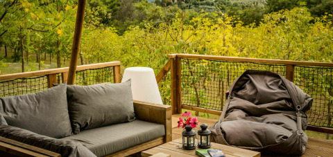 Zona de descanso exterior en una terraza de tienda safari con sofá, puf y faroles, rodeada de naturaleza.