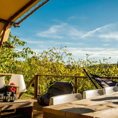 Zona de estar exterior junto a la tienda Desert Lodge con muebles de madera y vistas verdes en Camping Pian di Boccio.