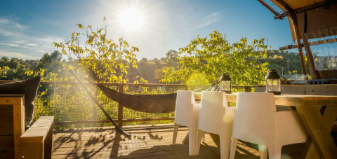 Terraza soleada con hamaca, sillas blancas y vista a los árboles en Desert Lodge en Camping Pian di Boccio.