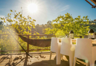 Terraza soleada con hamaca, sillas blancas y vista a los árboles en Desert Lodge en Camping Pian di Boccio.