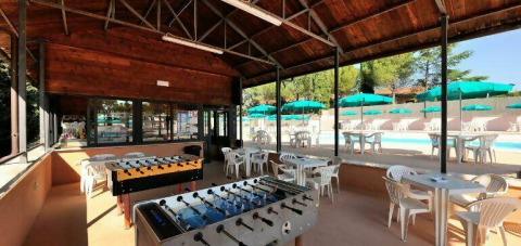 Espace couvert avec baby-foot et tables, vue sur piscine et parasols au Camping Pian di Boccio en Ombrie, Italie.