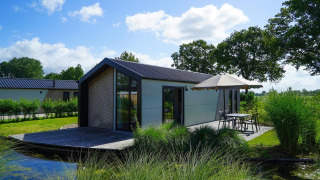 Lodge moderne au bord d’un lac avec terrasse et parasol, entouré de verdure et arbres.