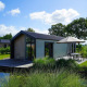 Moderne lodge ved søen med terrasse, parasol og have, omgivet af natur under en blå himmel.