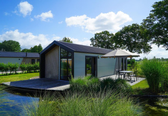 Moderne Lodge am See mit Terrasse und Sonnenschirm, eingebettet in grüne Natur und Bäume.