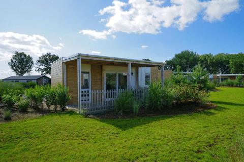 Un lodge moderne nommé Cosy Unique avec terrasse, entouré de pelouse et d’arbres sous un ciel bleu et ensoleillé.
