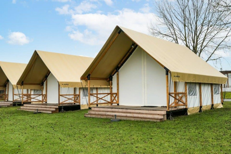 Safari glamping tents with beige canvas roofs and wood decks, set on green grass under a blue sky.