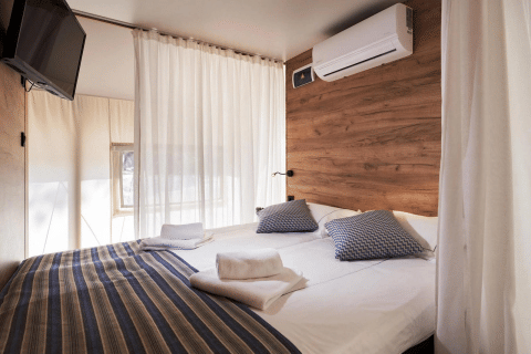 Moderne glampingtent slaapkamer met tweepersoonsbed, airco, televisie en houten accentmuur.