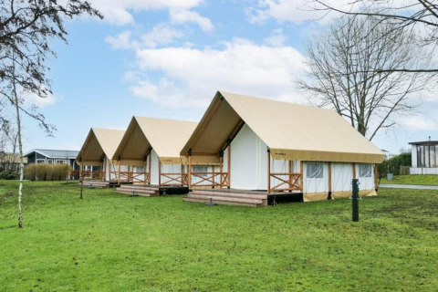 Drei luxuriöse Safarizelte auf einer grünen Wiese, umgeben von Bäumen und modernen Gebäuden im Hintergrund.