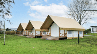 Drei luxuriöse Safarizelte auf einer grünen Wiese, umgeben von Bäumen und modernen Gebäuden im Hintergrund.