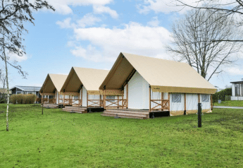 Trois tentes de glamping safari sur une pelouse verte avec arbres, ciel bleu et structures modernes autour.