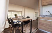 Dining area with a modern wooden table and black chairs inside a glamping tent, next to a kitchenette.