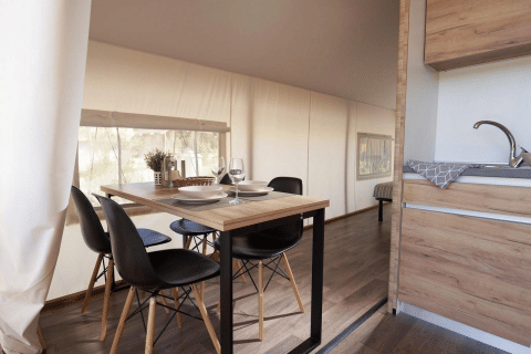 Espace repas avec table en bois moderne et chaises noires dans une tente glamping, près d’une kitchenette.
