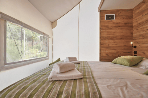Glampingtent bedroom with striped bedding, wood accent wall, towels on bed, and nature views outside.