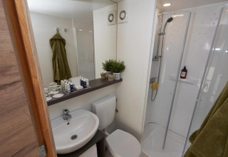Baño moderno y compacto en alojamiento glamping con ducha, inodoro, lavabo y toallas verdes visibles.