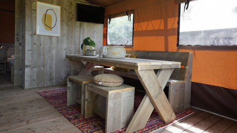 Interior de Glampingtent Desert con mesa de madera, bancos, alfombra colorida y cortinas de ventana naranjas.