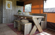 Indretning i Glampingtent Desert med træbord, bænke, farverig tæppe og vinduer med orange gardiner.