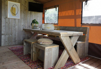 Interno della Glampingtent Desert con tavolo in legno, panche, tappeto colorato e tende arancioni alle finestre.