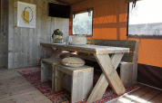 Intérieur de la Glampingtent Desert avec table en bois, bancs, tapis coloré et rideaux de fenêtre orange.