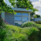 Et hyggeligt Pavilion-lodge med store vinduer, omgivet af grønne buske og farverige blomster i sollys.