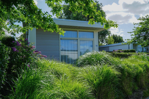 Gemütliche Pavilion-Lodge mit großen Fenstern, umgeben von grünen Büschen und Blumen an einem sonnigen Tag.